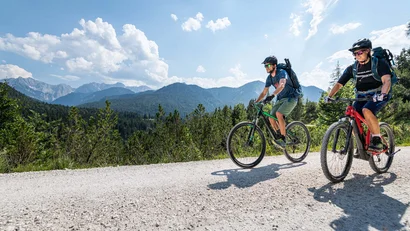 Two mountain bikers riding on a trail with mountains in the background on a sunny day.
