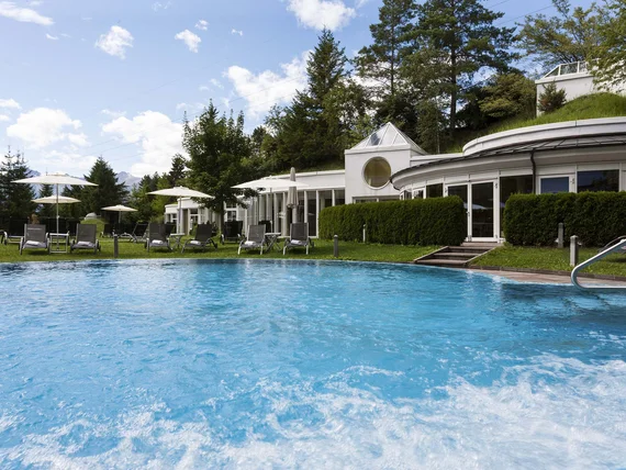 Pool with loungers and umbrellas in front of a hotel surrounded by greenery