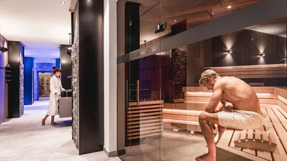 Man in towel sitting in bio sauna, woman in bathrobe at water station