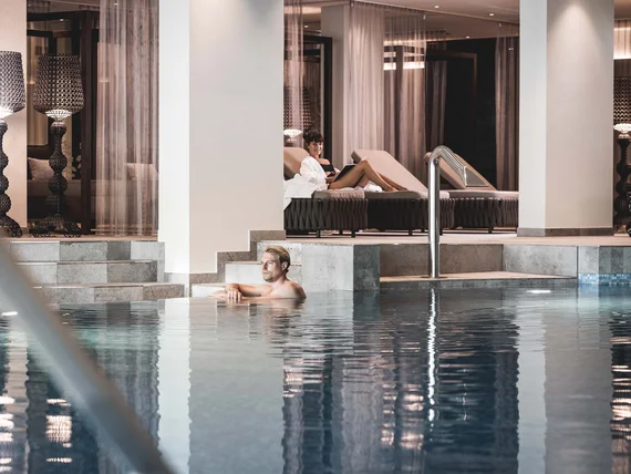 Relaxing man in indoor pool with woman on lounge chair in background